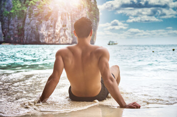 Back of young man sitting on beach on edge of the ocean