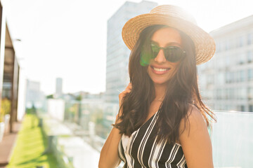 Close up portrait of fashionable stylish gilt with dark wavy hair wearing hat, sunglasses and summer t-shirt looking to camber Ian sunlight on city background 