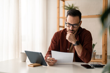 Serious millennial man entrepreneur looking at the paperwork taking notes while using computer digital technology, sitting at table in a home office during the day.