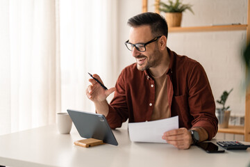 Smiling business person watching training webinar or having a video conference on digital tablet, working from home office, holding a pen taking notes while using digital tablet, sitting at the table.