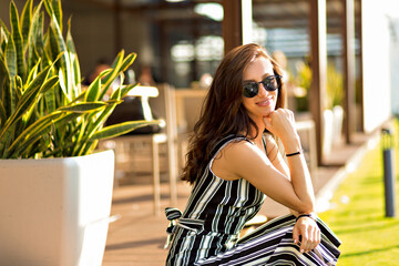 Close up outdoor photo of beautiful woman with dark hair wearing summer suit and sunglasses sitting on summer terrace with city view in sunny warm day