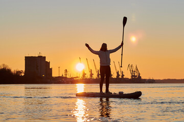 Silhouette of middle-aged woman on stand up paddle boards on a Danube river at winter sunrise against the backdrop of port cranes