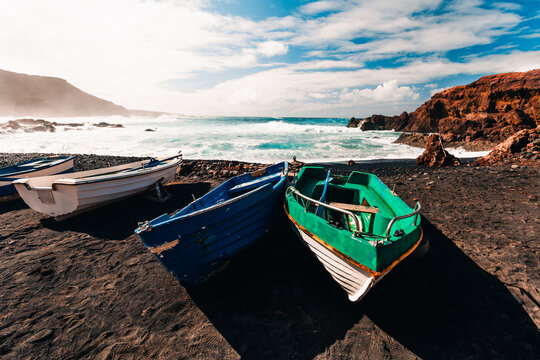 Landscape Photo of colorful boats (vessels) on volcanic black sand beach with beautiful waves in ocean on background - Lanzarote Island. Landscape shot of boats on the shore.