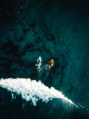Two surfers floating on surfboard in tropical ocean with big and powerful wave behind them. Aerial top view of surfers in crystal blue ocean - Vertical photo.