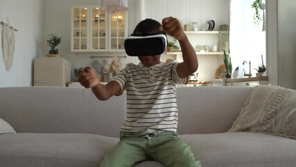 Little African American boy child wearing VR headset sitting on sofa at home playing enjoyable car racing game. Small kid spending leisure time in virtual reality. Children and 3d gaming
