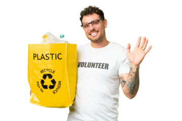 Brazilian man holding a bag full of plastic bottles to recycle over isolated chroma key background saluting with hand with happy expression