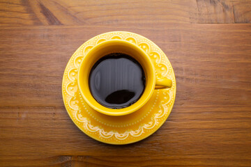 Uma xícara amarela de café preto com um pires amarelo sobre uma mesa de madeira.