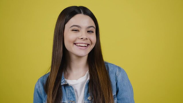 Lovely teen girl smiling a toothy smile, blinking looking at camera, isolated over yellow background