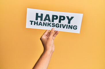 Hand of caucasian man holding paper with happy thanksgiving message over isolated yellow background
