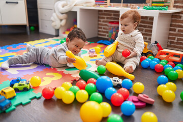 Two adorable babies playing with balls and bowling pin sitting on floor at kindergarten
