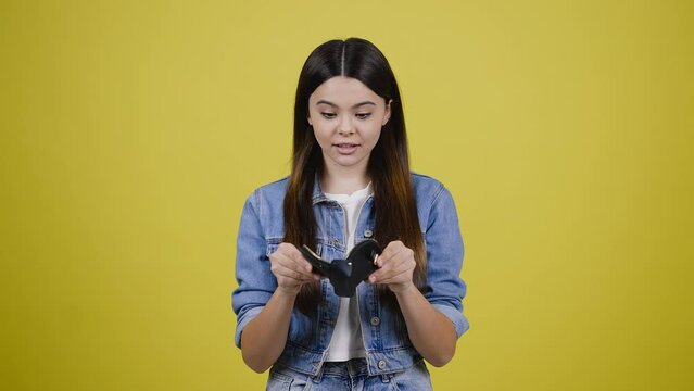 Smiling pretty teenage girl expressing sadness noticing her leather wallet empty, yellow background