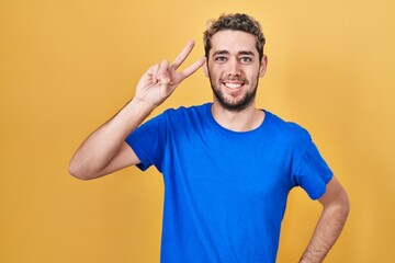 Hispanic man with beard standing over yellow background smiling looking to the camera showing fingers doing victory sign. number two.