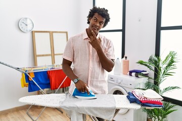African man with curly hair ironing clothes at home with hand on chin thinking about question,...