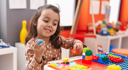 Adorable hispanic girl playing with construction blocks sitting on table at kindergarten