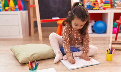 Adorable hispanic girl preschool student sitting on floor drawing on notebook at kindergarten