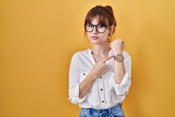 Young beautiful woman wearing casual shirt over yellow background in hurry pointing to watch time,...