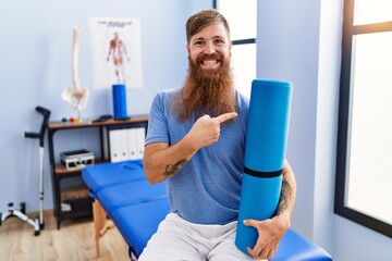 Redhead man with long beard holding yoga mat at rehabilitation clinic smiling happy pointing with...