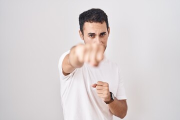 Handsome hispanic man standing over white background punching fist to fight, aggressive and angry...
