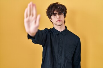 Young man wearing glasses over yellow background doing stop sing with palm of the hand. warning...