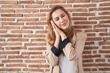 Beautiful blonde woman standing over bricks wall sleeping tired dreaming and posing with hands together while smiling with closed eyes.