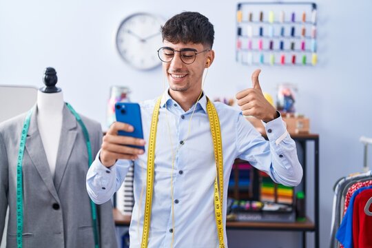 Young hispanic man dressmaker designer using smartphone smiling happy and positive, thumb up doing excellent and approval sign