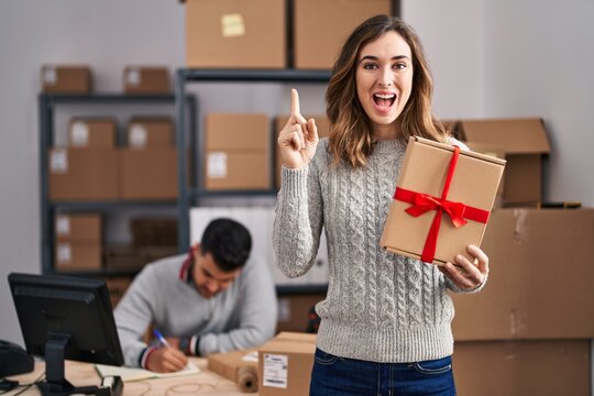 Young hispanic people working at small business ecommerce holding gift smiling with an idea or question pointing finger with happy face, number one