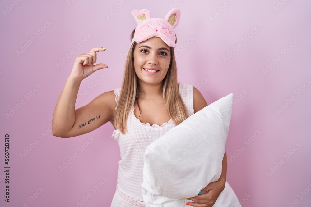 Canvas Prints Young blonde woman wearing pyjama hugging pillow smiling and confident gesturing with hand doing small size sign with fingers looking and the camera. measure concept.