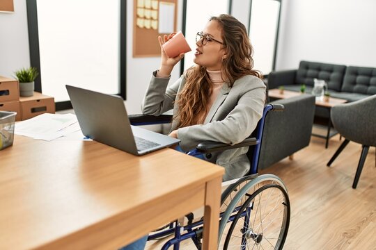 Young beautiful hispanic woman business worker drinking coffee sitting on wheelchair at office