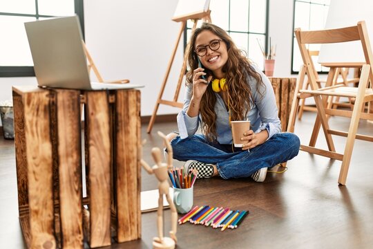 Young beautiful hispanic woman artist talking on smartphone drinking coffee at art studio