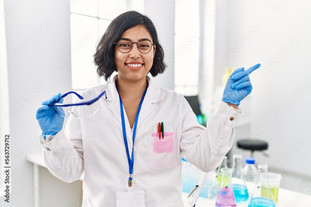 Poster Young hispanic woman working at scientist laboratory holding safety glasses smiling happy pointing with hand and finger to the side