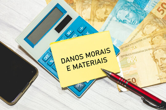 As palavras Danos morais e materiais escrita em um pedaço de papel que está sobre uma calculadora. Notas do Real Brasileiro na composição. Economia brasileira.