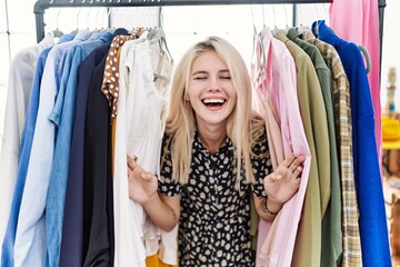Naklejka na ściany i meble Young blonde woman searching clothes on clothing rack smiling and laughing hard out loud because funny crazy joke.