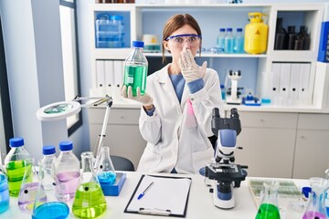 Young brunette woman working at scientist laboratory covering mouth with hand, shocked and afraid...