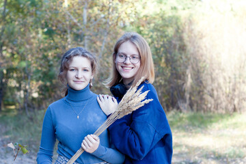 Fototapeta premium Two young beautiful smiling teenage girls in nature. Happy sisters, dressed in blue sweaters, with bouquet of dried flowers in their hands. Walk in autumn park. Friendship, love, teenagers