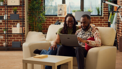 Boyfriend and girlfriend watching video on laptop together, sitting on couch and relaxing at home. Young partners in relationship enjoying leisure activity online, stay home laughing. Tripod shot.