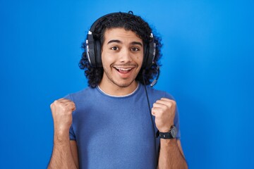 Hispanic man with curly hair listening to music using headphones celebrating surprised and amazed for success with arms raised and open eyes. winner concept.