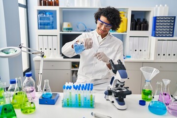 Young hispanic man wearing scientist uniform pouring liquid on test tube at laboratory