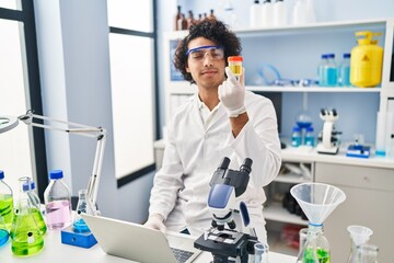 Young hispanic man wearing scientist uniform using laptop analysing urine test tube at laboratory