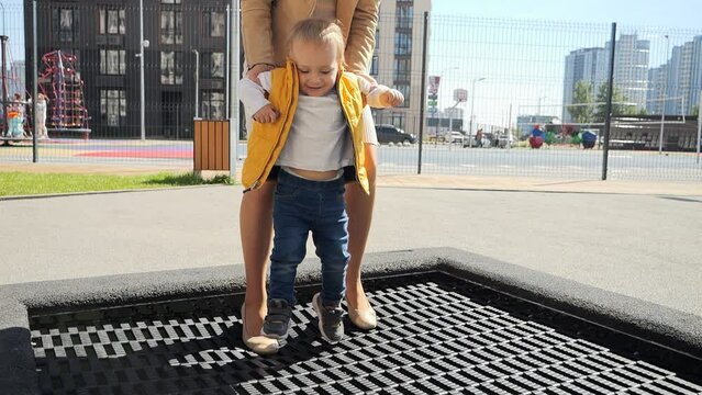Slow motion of young mother holding her baby son jumping on outdoor trampoline at playground. Happy parenting, family having time together, kids and parents outdoors