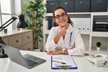 Young hispanic woman wearing doctor uniform and stethoscope looking confident at the camera smiling with crossed arms and hand raised on chin. thinking positive.