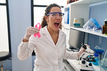 Young hispanic woman working at scientist laboratory looking for breast cancer cure angry and mad screaming frustrated and furious, shouting with anger. rage and aggressive concept.