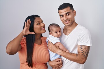 Young hispanic couple with baby standing together over isolated background smiling with hand over ear listening an hearing to rumor or gossip. deafness concept.