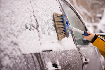 A boy with a brush in his hands cleans the car from snow. The car is covered of snow. The car prepares for a trip after a snowfall. Safety first
