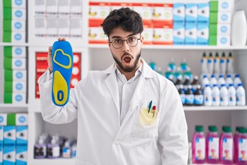 Hispanic man with beard working at pharmacy drugstore holding insole scared and amazed with open mouth for surprise, disbelief face