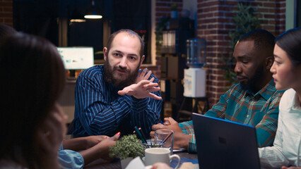 Diverse coworkers brainstorming in business meeting, discussing presentation, team communication, executive zoom in. Employees planning sales strategy in coworking space at night time