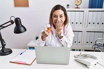 Young hispanic woman wearing doctor uniform holding pills at the clinic laughing and embarrassed...