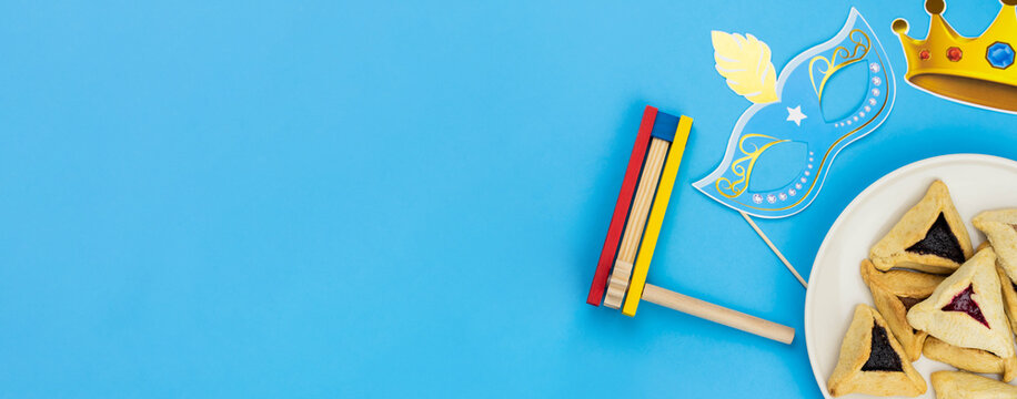 Jewish holiday Purim celebration concept. Traditional hamantasch or hamantaschen Haman's ears cookies, carnival mask, spin gragger (ratchet) noisemaker, and party decor on blue background.