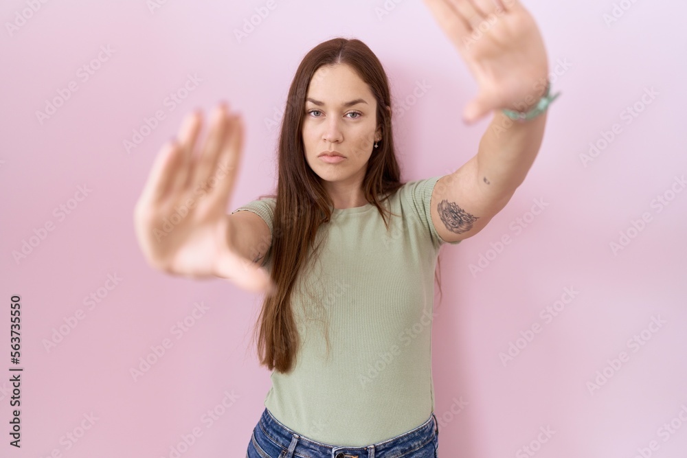 Poster Beautiful brunette woman standing over pink background doing frame using hands palms and fingers, camera perspective