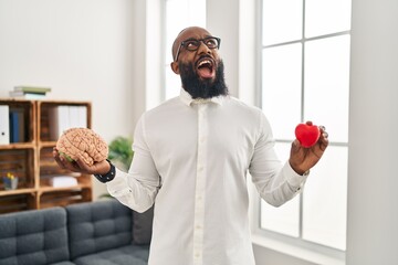 African american man working at therapy office holding brain and heart angry and mad screaming...