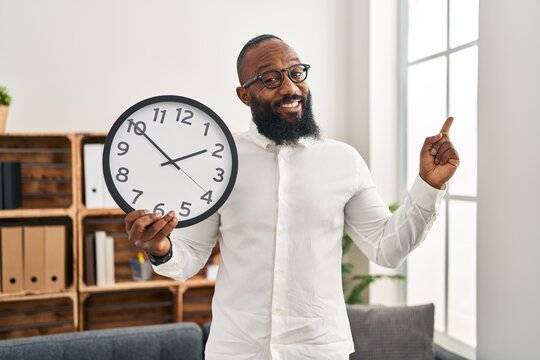 African american man holding big clock at the office smiling happy pointing with hand and finger to the side